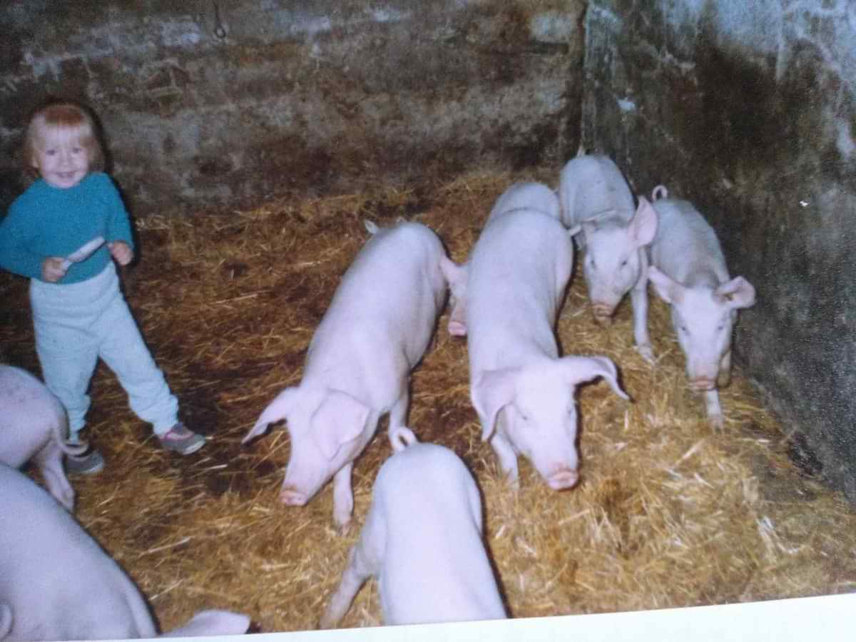 A young Emilia Mikulewicz on her family farm in 1990s Poland, standing in a straw-filled pen among piglets — a glimpse into her early life rooted in hands-on agricultural experience during a time of post-communist transition.